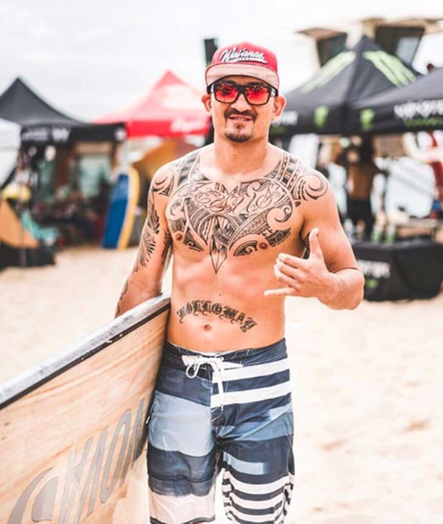 Tatuado Bomber Team Surfer dando un shaka mientras sostiene su tabla en Nicaragua